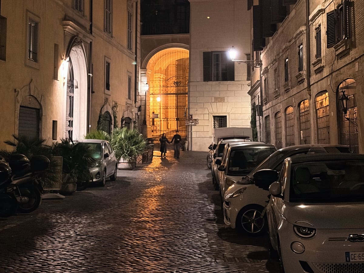 Narrow cobblestone street at night with parked cars and motorbikes, flanked by tall, shuttered buildings. A glowing archway illuminates two distant pedestrians under warm streetlights.